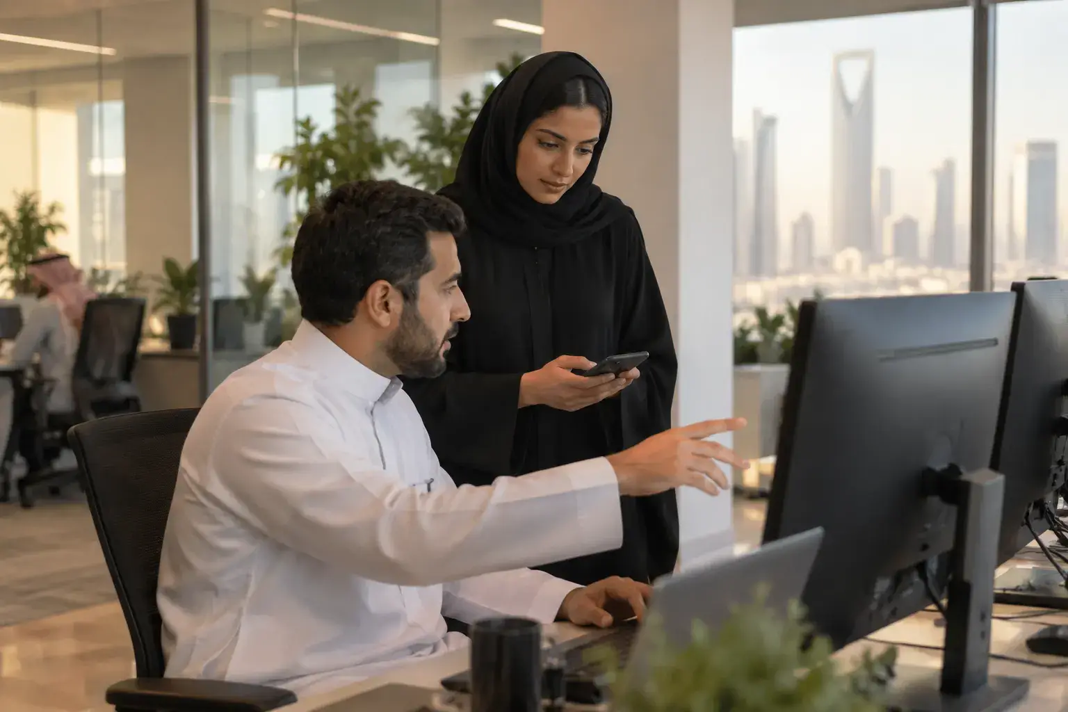 An IT support engineer in a thobe at a Riyadh head office helps a colleague in an abaya who holds her phone, two monitors angled away.