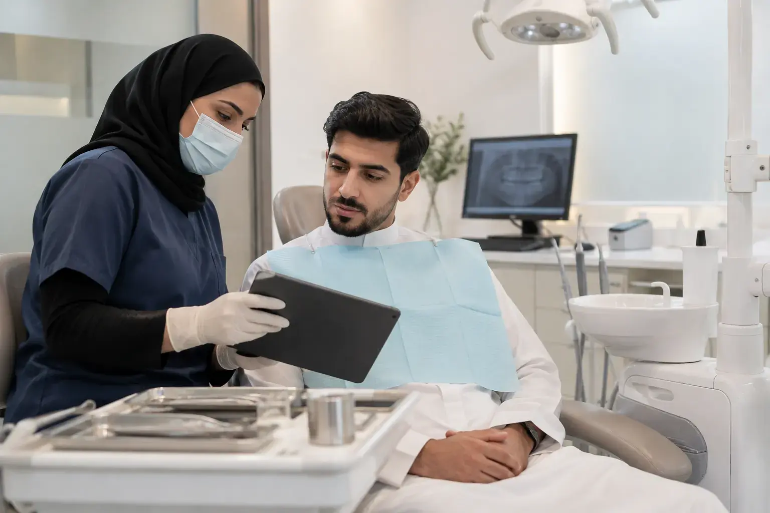 A Saudi dentist in a hijab and mask shows a young patient in a thobe something on a tablet in a Riyadh dental treatment room.