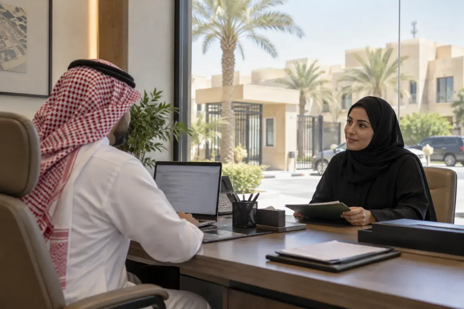 A Saudi leasing manager in a white thobe and red shemagh at a laptop in a villa compound’s leasing office in Al Malqa, Riyadh, talking with a woman in a black abaya holding a folder.