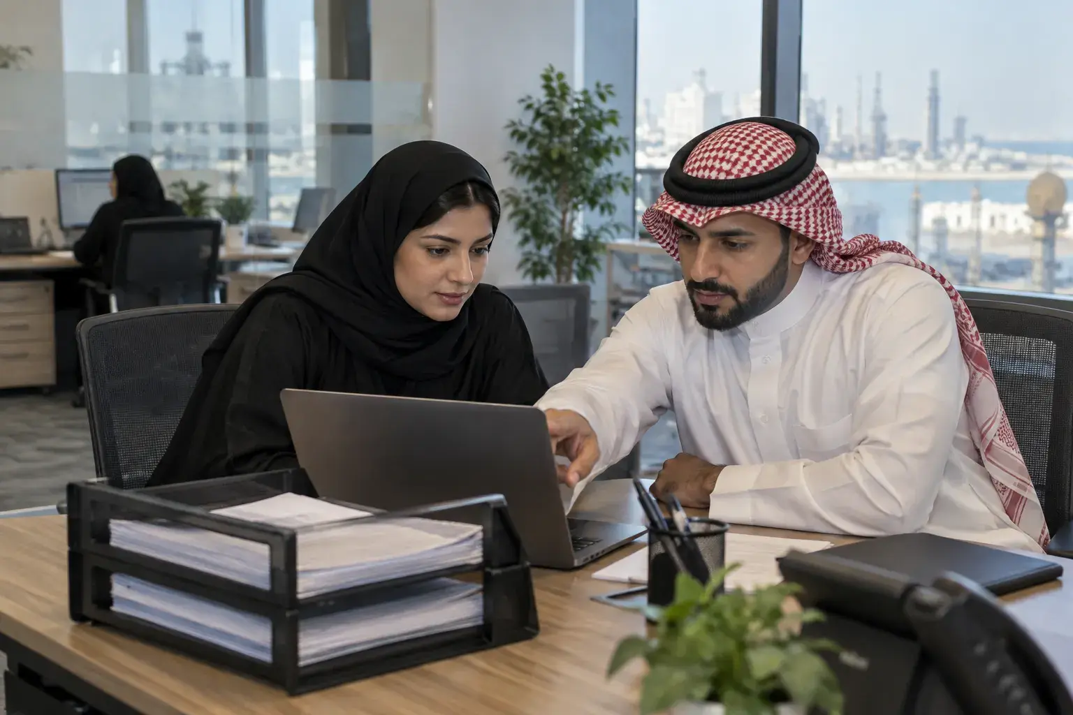 A Saudi HR officer in an abaya and an accountant in a thobe and shemagh review a laptop together in a Dammam office, with printed payslips in a tray.