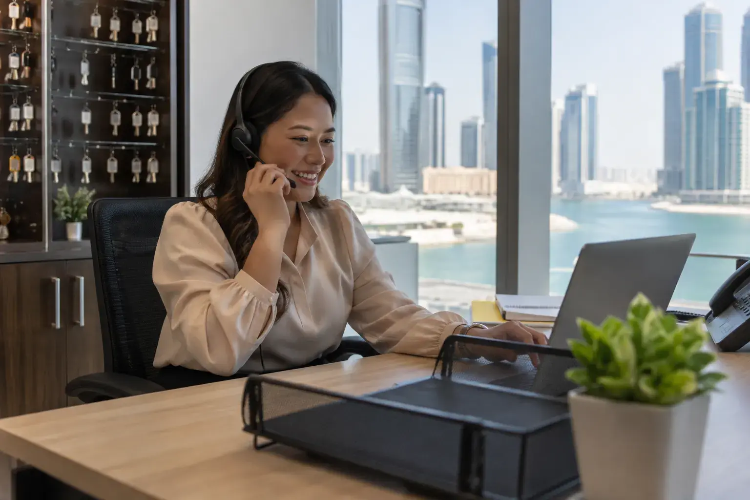 A leasing coordinator on a headset at a tidy desk in an Al Reem Island office in Abu Dhabi, a key cabinet behind her and the water outside.