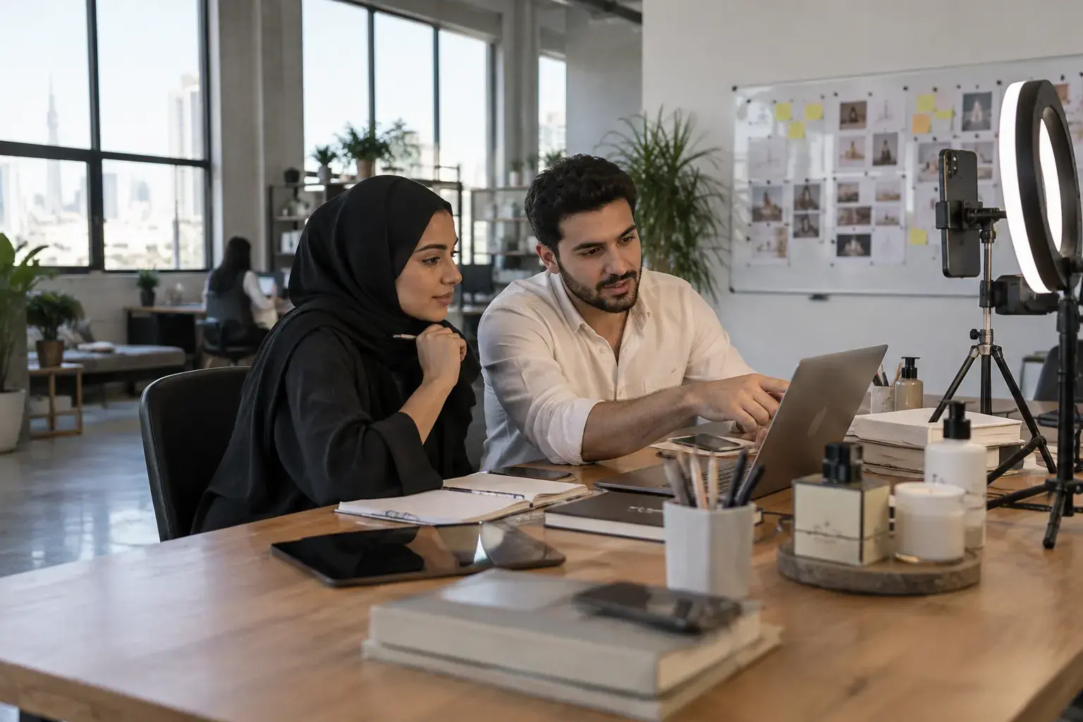 A content lead and a designer review posts on a laptop at a social media agency studio in Al Quoz, Dubai, beside a ring light and a phone on a tripod.