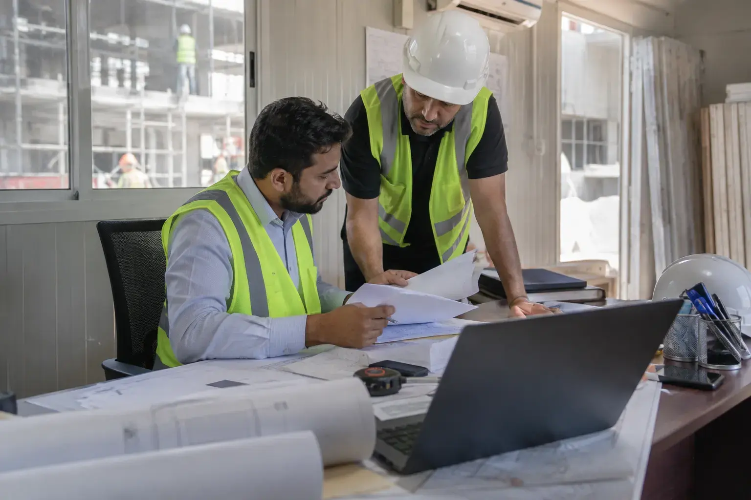 A quantity surveyor and a site manager in safety vests check supplier delivery notes at a desk in a portacabin site office in Al Quoz, a building under fit-out outside.
